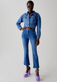 Giacca di jeans tagliata e jeans a zampa in denim blu, abbinati a tacchi a piattaforma viola e occhiali da sole scuri. Texture liscia, vestibilità classica.