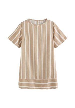 Kobieta ubrana w beżowo-białą sukienkę w pionowe paski z krótkim rękawem i kieszeniami, złote kolczyki i okulary przeciwsłoneczne, stojąca przy białej ścianie i balustradzie.