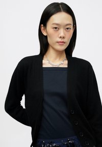 Cardigã preto com uma textura suave, de frente aberta, e seis botões. Usado sobre uma blusa azul-marinho, complementado por um colar de prata com detalhes redondos.