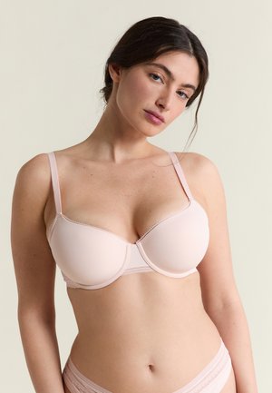 Vrouw met lichte huid en donker haar, gekleed in een lichtroze beha en bijpassend ondergoed tegen een effen beige achtergrond.