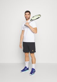 Witte polo, zwarte sportshorts, blauwgroene sneakers, met een tennisracket in de hand, staand tegen een effen witte achtergrond.