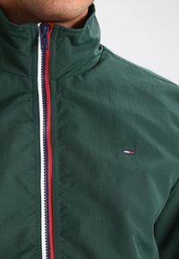 Muž nosící tmavě zelenou bundu na zip s červeno-bílo-modrým proužkovaným zipem a malým vyšívaným logem na hrudi.