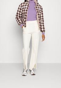 Brązowa i biała kurtka puchowa w kratę z wysokim kołnierzem, fioletowy golf, białe spodnie z bocznymi rozporkami oraz wzorzyste sneakersy.