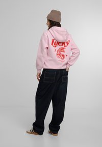 Sudadera rosa con el texto impreso en rojo "Lucky" y diseño de cereza en la parte de atrás, combinada con jeans oscuros anchos y un gorro beige.