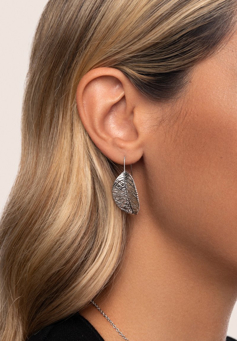 Pendientes en forma de hoja de plata con detalle texturizado, colgando de un fino gancho, usados sobre un fondo neutro.
