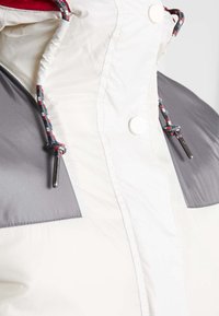 Gros plan d'une veste blanche et grise avec des cordons multicolores et des boutons-pression blancs sur le col.