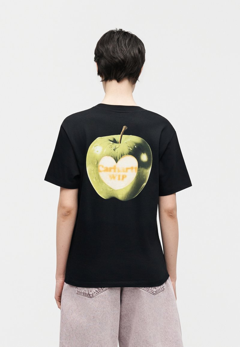 Person mit kurzen dunklen Haaren, die ein schwarzes T-Shirt mit einem großen grünen Apfel und einem herzförmigen Lichtbereich trägt, auf dem auf der Rückseite "Carhartt WIP" steht.