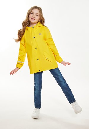 Chaqueta de lluvia impermeable amarilla con capucha, botones a presión y bolsillos delanteros. Llevada con jeans azules y zapatillas blancas. Textura suave.