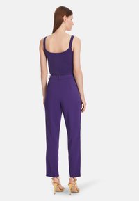 Purple fitted top with wide straps paired with high-waisted purple trousers. Model wears gold high-heeled sandals. Smooth, sleek texture.