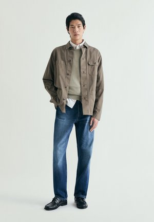 Hombre joven de pie que lleva una chaqueta marrón sobre un suéter beige, camisa blanca, vaqueros azules y zapatos negros sobre un fondo claro y liso.