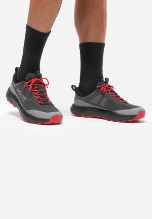 Jambes portant des chaussettes noires et des chaussures de trail gris et rouge avec des lacets rouges sur un fond blanc.