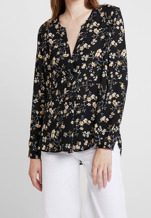 Black long-sleeve blouse with small yellow and white floral print worn with white pants, shown from shoulders to hips.