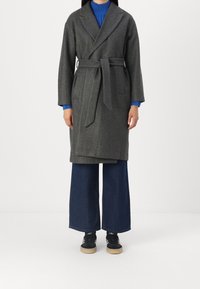 Grijze wollen mantel met een strikceintuur, lange mouwen en een sjaalkraag, gedragen over donkerblauwe denim broek met wijde pijpen en zwarte sneakers.