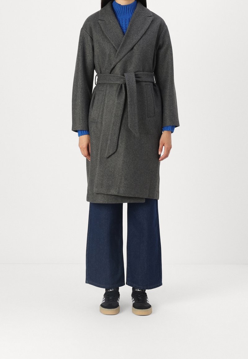 Grijze wollen mantel met een strikceintuur, lange mouwen en een sjaalkraag, gedragen over donkerblauwe denim broek met wijde pijpen en zwarte sneakers.