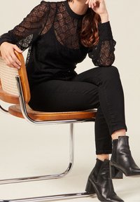 Blusa nera in pizzo con motivi geometrici e maniche lunghe, abbinata a jeans neri slim e stivaletti con tacco neri, seduta su una sedia.