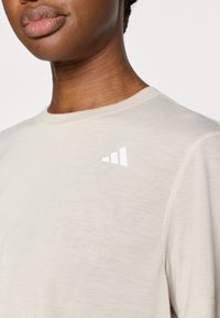 Ανοιχτό μπεζ μπλουζάκι με κοντά μανίκια, στρογγυλή λαιμόκοψη, απαλό ύφασμα, με λευκό τριγωνικό λογότυπο στον αριστερό ώμο.