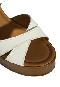 Sandale en cuir synthétique blanc avec des lanières croisées, une semelle intérieure rembourrée marron et une semelle en bois épaisse. Texture lisse avec des détails de couture.