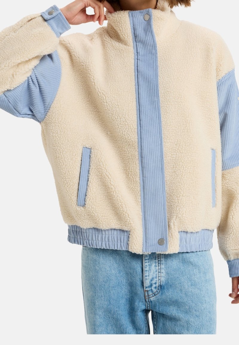 Personne portant une veste en polaire crème avec des accents en tissu côtelé bleu clair et un jean en denim bleu clair, main près du visage.