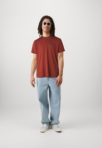 Červené tričko s okrúhlym výstrihom a zeleným logom, skombinované so svetlomodrými džínsami a bielymi topánkami. Model má na sebe slnečné okuliare a stojí na neutrálnom pozadí.