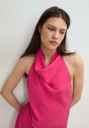 Mlada ženska z dolgimi temnimi lasmi, oblečena v svetlo roza zgornji del brez rokavov z nagubanim dizajnom, stoji ob navadni svetli steni, gleda navzdol in v stran.