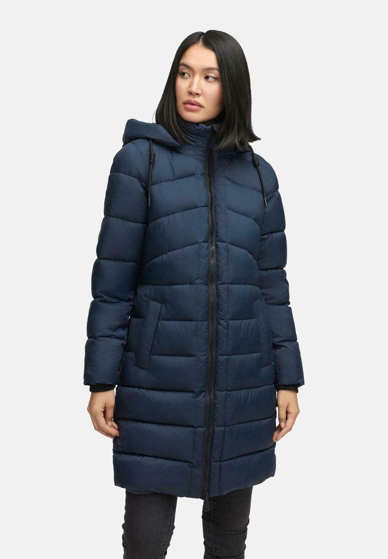 Mujer vistiendo un abrigo de invierno acolchado con capucha en azul marino, con cremallera frontal y bolsillos, de pie con las manos relajadas a los lados, mirando hacia la izquierda.