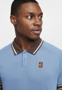 Mann iført lyseblå perforert polo skjorte med brun krage og Nike-logo, som ser mot høyre foran en ensfarget bakgrunn.