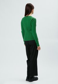 Grüner gerippter Pullover mit langen Ärmeln und enganliegendem Saum, kombiniert mit schwarzen weit geschnittenen Hosen und klobigen schwarzen Stiefeln.