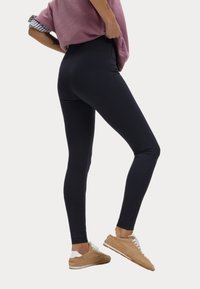 Zwarte high-waisted legging van rekbare stof, met een gladde textuur en een slanke pasvorm, gecombineerd met een losse roze top en beige sneakers.