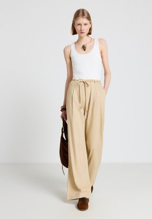 Donna con top bianco senza maniche, pantaloni beige a gamba larga con coulisse, scarpe marroni e che tiene una borsa marrone su sfondo bianco uniforme.