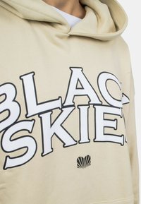 Béžová mikina s kapucňou s veľkým čiernym nápisom "BLACK" a "SKIES", pod ktorým je malá grafika mušle. Mäkko textúrovaná látka.