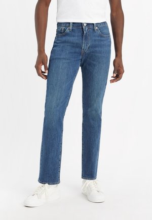 Man die een blauwe rechte spijkerbroek, witte sneakers en een wit T-shirt draagt tegen een effen witte achtergrond.