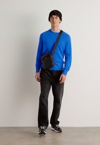 Maglione blu brillante, jeans neri, borsa a tracolla nera e scarpe da ginnastica nere. Modella in piedi di fronte a uno sfondo bianco semplice.