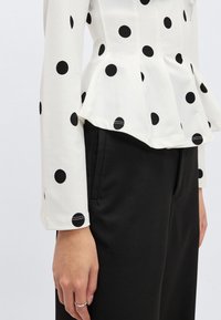 Hvitt peplum-topp med svarte polkadotter, lange ermer og glatt stofftekstur, kombinert med svarte bukser. Naturlig håndposisjon synlig.