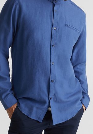 Mężczyzna w niebieskiej, długorękawowej koszuli zapinanej na guziki z kieszenią na piersi, z rękami w kieszeniach ciemnych spodni, na jednolitym jasnym tle.