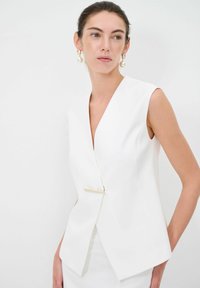 Gilet bianco senza maniche con scollatura a V profonda e un elegante dettaglio in metallo dorato in vita. Texture liscia e design sartoriale.