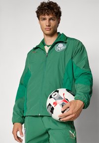 Grüne sportliche Jacke mit Reißverschluss, Logo und kontrastierenden Farbpaneelen. Das Model hält einen schwarz-weißen Fußball. Weicher, leichter Stoff.