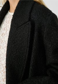 Abrigo negro texturizado con solapa y un corte holgado, fabricado en un suave y aterciopelado tejido. La capa interior presenta un ligero patrón de encaje.