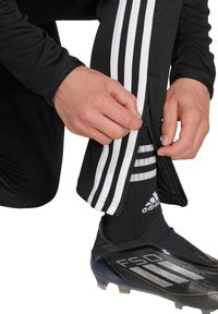 Schwarze Sporthose mit weißen Streifen, Reißverschlüssen, getragen mit schwarzen Fußballschuhen und Socken, mit einem Logo und gestreiften Mustern.