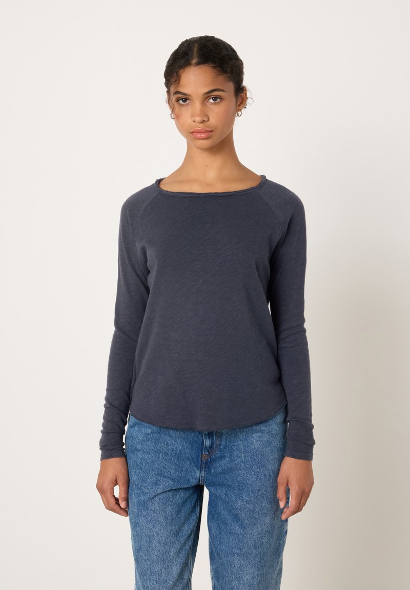 Moteris su garbanotais plaukais, dėvinti tamsią ilgomis rankovėmis palaidinę ir mėlynas džinsines kelnes, stovinti priešais šviesų, paprastą foną.