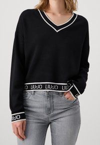 Pull-over noir court avec encolure en V, présentant des accents blancs et une bande de marque à la taille et aux poignets. Le matériau semble doux et tricoté.