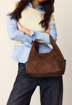 Ženska, oblečena v modro črtasto srajco in kremasto jopico, drži veliko rjavo usnjeno torbico z zlatim okovjem ob pasu.