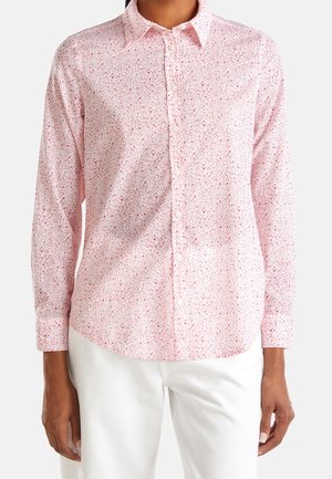 Henkilö, jolla on päällään pitkähihainen, napitettava vaaleanpunainen paitapusero, jossa on pieni kukkakuosi, sekä valkoiset housut, seisomassa yksiväristä taustaa vasten.