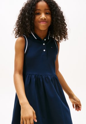 Junges Mädchen mit lockigem Haar, das ein ärmelloses marineblaues Kleid mit geknöpftem Kragen trägt und vor einem schlichten weißen Hintergrund steht.