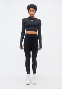 Άτομο που στέκεται κρατώντας αλτήρες 1 κιλού σε κάθε χέρι, φορώντας μαύρο crop top με μακριά μανίκια, μαύρο κολάν, λευκές κάλτσες και λευκά αθλητικά παπούτσια.