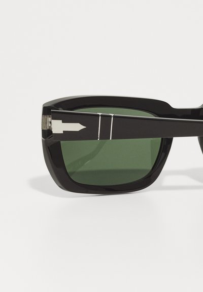 Occhiali da sole neri con una montatura spessa e angolare, lenti verdi e un dettaglio a freccia argentato sulla stanghetta. La montatura ha una finitura lucida.