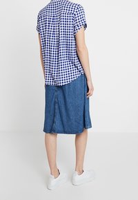 Chemise à manches courtes à carreaux bleus et blancs, assortie à une jupe en denim longueur genou et des baskets blanches, vue de dos.