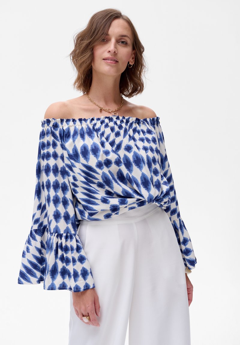 Bluzka na ramiączkach w białym materiale z niebieskimi wzorami liści. Posiada dzwonowe rękawy iwiązany przód. Połączona z białymi spodniami.