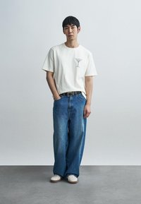 Άσπρο βαμβακερό μπλουζάκι με μικρό μαύρο σχέδιο δέντρου; χαλαρό μπλε τζιν; άσπρα αθλητικά παπούτσια; μαύρη ζώνη; στέκεται μπροστά σε απλό φόντο.