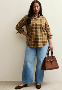 Camisa de cuadros con botones en tonos amarillos y marrones, combinada con jeans de pierna ancha en azul claro. Los accesorios incluyen un bolso marrón y zapatos planos oscuros.