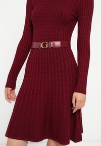 Donna che indossa un abito lungo in maglia bordeaux con motivi a rombi e a treccia, stretto in vita con una cintura abbinata che presenta una fibbia dorata a forma di "G".